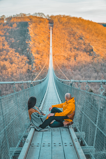 Photo of young couple sitting on a hanging bridge in Germany during the summer day, suspension rope bridge. Young female sitting with her boyfriend outdoor and admiring view Travel Lifestyle concept with rope bridge background and sunset. Young woman and man hanging rope bridge with sky as background. Leisure extreme activity at autumn day. Beautiful tourist on an excursion.