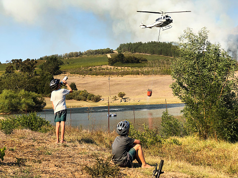 Two Caucasian Cycling boys watching a rescue firefighting helicopter filling a water bucket from a dam lake with the fire cloud of smoke behind near Stellenbosch South Africa