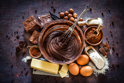 Top view of dark kitchen table filled with ingredients for preparing homemade chocolate brownies. The composition is arranged at the center of the frame. A large mixing bowl filled with chocolate spread mix and a wire whisk is at the center and all around it are other ingredients like butter, powdered cocoa, sugar, flour, eggs, chocolate bars salt and hazelnuts. Predominant color is brown. Low key DSRL studio photo taken with Canon EOS 5D Mk II and Canon EF 100mm f/2.8L Macro IS USM.
