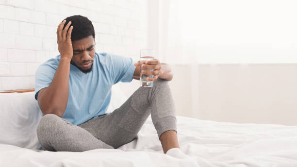 Frustrated young man touching his head and keeping eyes closed Feeling unwell, hangover. Frustrated young african-american man touching his head, holding glass of water in bed, copy space drought stock pictures, royalty-free photos & images