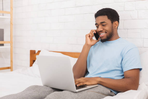 hombre sonriente, hablando por teléfono y el uso de laptop en la cama - working bed smart phone bedroom fotografías e imágenes de stock