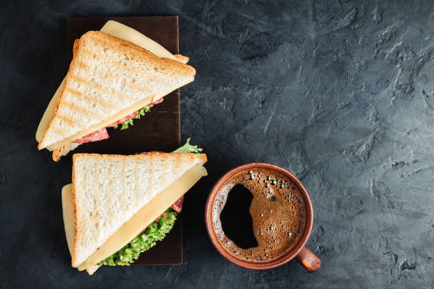 tazza con caffè caldo e sandwich con toast alla griglia, salsiccia di salame, lattuga di insalata e formaggio su sfondo scuro, vista dall'alto - cheese sandwich foto e immagini stock