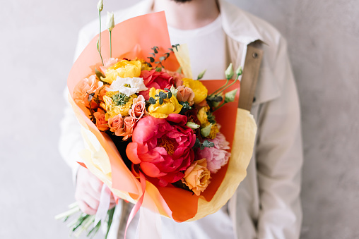 very nice young man holding a beautiful blossoming flower bouquet of fresh peony, tulips, roses, eustoma in yellow, coral and pink colors on the grey wall background