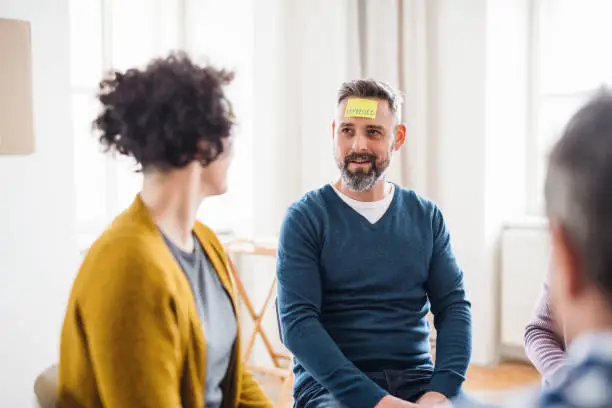 Photo of Men and women sitting in a circle during group therapy, adhesive notes on forehead.