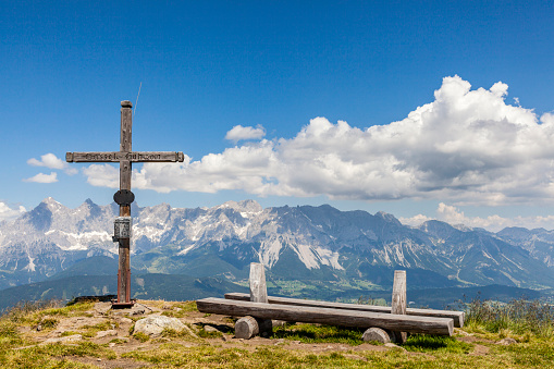 Wide panoramic landscape view from summit of mountain Gasselhoehe with cross and wooden bench to mountain range Dachstein in Styria, Austria