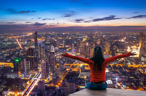 Rear view of female traveler with raised hands standing on the top of a tower and looking at the  urban city with sunset sky. Woman happy enjoying the city view from top of a city at twilight.