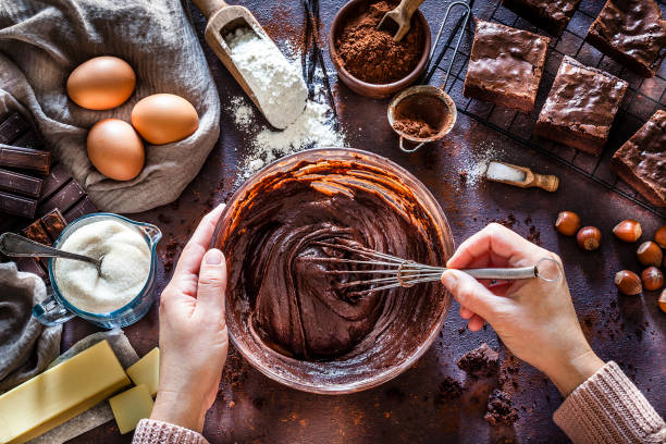 Chocolate brownie preparation on kitchen table Top view of dark kitchen table filled with ingredients for preparing homemade chocolate brownies. At the center of the frame is a large mixing bowl with chocolate spread mix and coming from the bottom of the frame are female hands holding the bowl and a wire whisk mixing the chocolate spread mix. At the top right are several prepared brownies on a cooling rack. Scattered on the table are other ingredients like butter, powdered cocoa, sugar, flour, eggs, chocolate bars salt and hazelnuts. Predominant color is brown. Low key DSRL studio photo taken with Canon EOS 5D Mk II and Canon EF 100mm f/2.8L Macro IS USM. wire whisk stock pictures, royalty-free photos & images