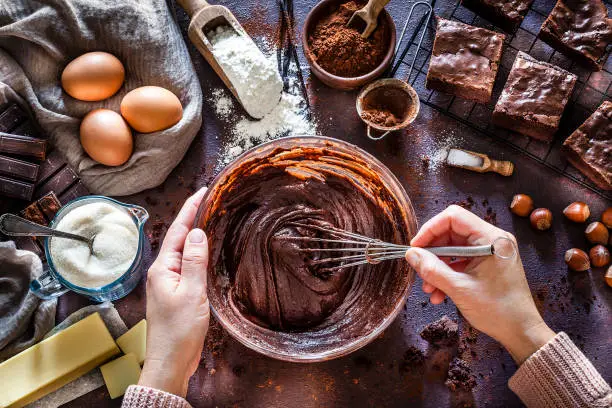 Top view of dark kitchen table filled with ingredients for preparing homemade chocolate brownies. At the center of the frame is a large mixing bowl with chocolate spread mix and coming from the bottom of the frame are female hands holding the bowl and a wire whisk mixing the chocolate spread mix. At the top right are several prepared brownies on a cooling rack. Scattered on the table are other ingredients like butter, powdered cocoa, sugar, flour, eggs, chocolate bars salt and hazelnuts. Predominant color is brown. Low key DSRL studio photo taken with Canon EOS 5D Mk II and Canon EF 100mm f/2.8L Macro IS USM.