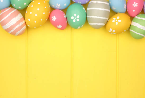 Colorful pastel Easter Egg top border against a yellow wood background. Above view with copy space.
