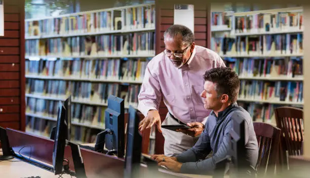Two multi-ethnic men in the library looking at a computer screen and conversing. A mature man in his 40s is sitting at the desktop PC and a senior African-American man in his 60s is standing beside him.