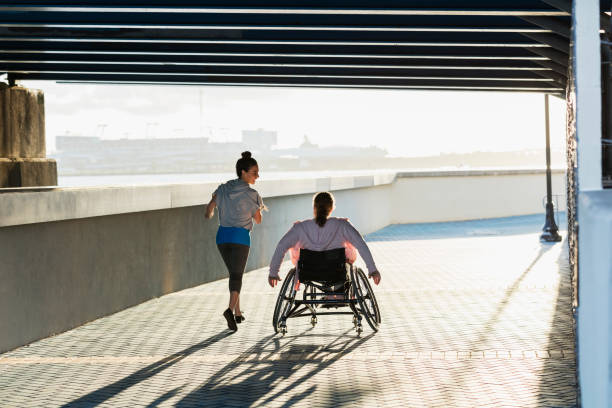 Young woman with spina bifida, Hispanic friend jogging Rear view of two friends exercising along a city waterfront. The young woman in the wheelchair has spina bifida. Her friend jogging beside her and talking is a mid adult Hispanic woman in her 30s. wheelchair stock pictures, royalty-free photos & images
