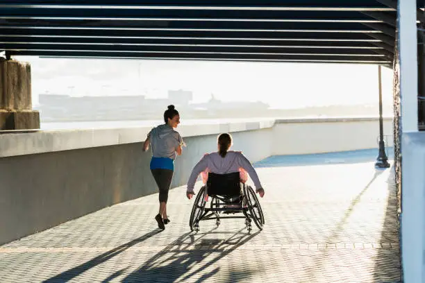 Rear view of two friends exercising along a city waterfront. The young woman in the wheelchair has spina bifida. Her friend jogging beside her and talking is a mid adult Hispanic woman in her 30s.