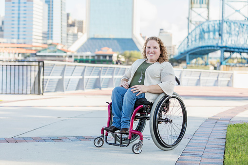 A young woman in her 20s in a self-propelled wheelchair, smiling at the camera.  She is out on her own in a city, travelling along the waterfront on a pedestrian walkway. She was born with spina bifida.