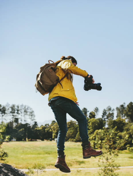 Explorer walking through tough terrain in the countryside holding a dslr camera. Rear view of a man wearing backpack jumping over a wooden log holding a digital camera in hand.