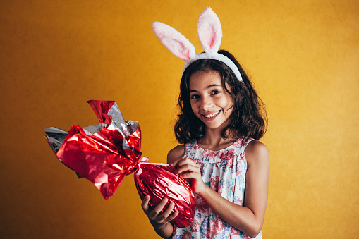 Cute little child wearing bunny ears on Easter day on color background. Girl holding chocolate easter egg