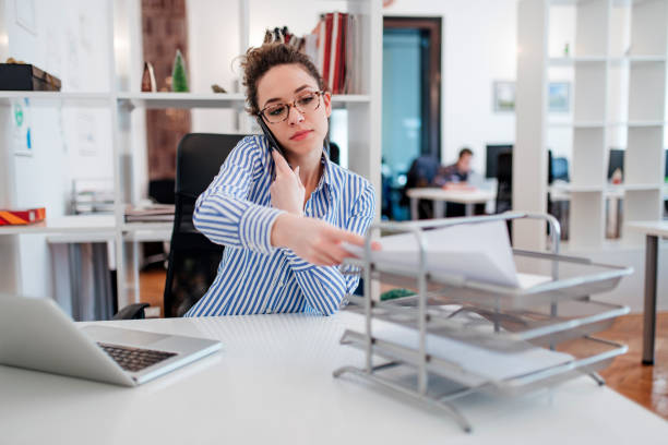 Young lady at desk doing paperwork and talking on smartphone. Young lady at desk doing paperwork and talking on smartphone. file clerk stock pictures, royalty-free photos & images