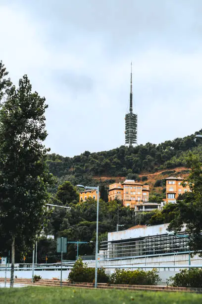 A vertical view of the tv tower "Torre de Collserola" in Barcelona, Spain on an overcast day with greenery and several residential buildings in the foreground and partly hidden road with poles around