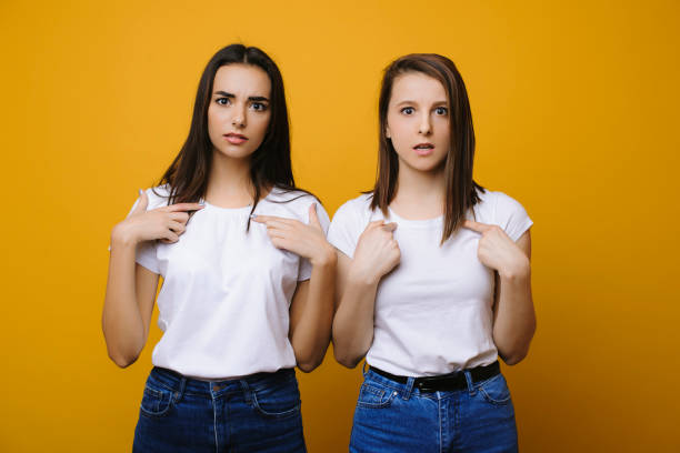 istock Two lovely young girl pointing on themselves perplex while looking into camera against yellow background. 1130573935