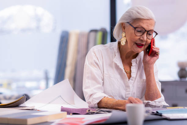 Enchanting well-groomed aging boss speaking on the phone in office. Telephone conversation. Enchanting well-groomed aging boss in white stylish shirt and trendy accessories busily speaking on the phone in office. thick rimmed spectacles stock pictures, royalty-free photos & images