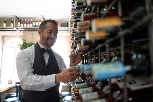 Friendly latin american waiter at a restaurant looking for a wine bottle on rack smiling very happy