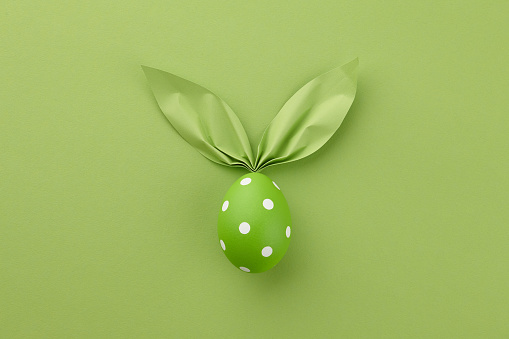 Green dotted Easter egg with paper bunny ears on spring green colored background. Holiday concept.