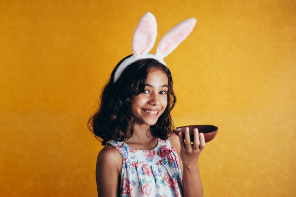 симпатичный маленький ребенок носить кролика уши на пасху день на цветном фоне. девушка ест шоколадное пасхальное яйцо - child easter flower little girls стоковые фото и изображения