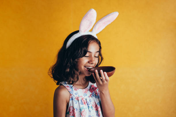 bambino carino che indossa orecchie da coniglietto il giorno di pasqua su sfondo a colori. ragazza che mangia l'uovo di pasqua al cioccolato - easter easter egg child chocolate foto e immagini stock