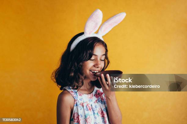 Cute Little Child Wearing Bunny Ears On Easter Day On Color Background Girl Eating Chocolate Easter Egg Stock Photo - Download Image Now