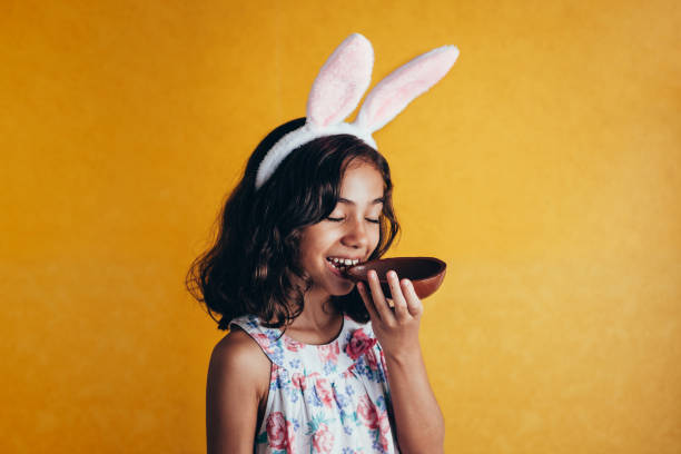 симпатичный маленький ребенок носить кролика уши на пасху день на цветном фоне. девушка ест шоколадное пасхальное яйцо - child easter flower little girls стоковые фото и изображения