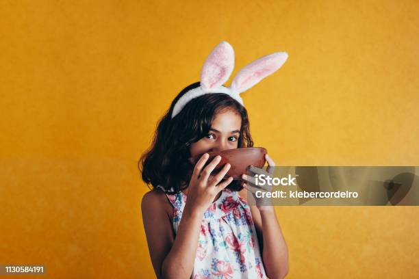 Cute Little Child Wearing Bunny Ears On Easter Day On Color Background Girl Eating Chocolate Easter Egg Stock Photo - Download Image Now