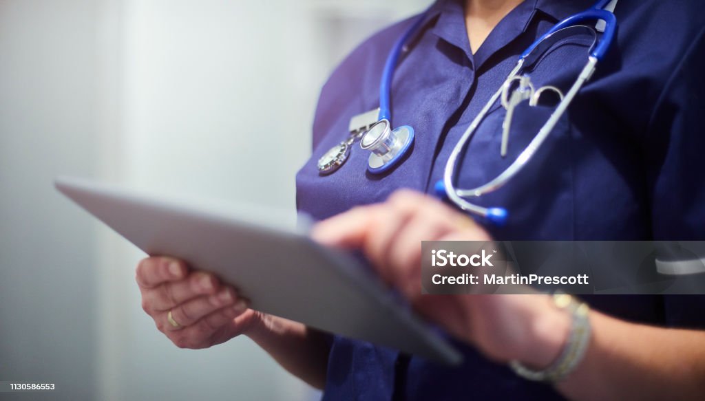 Doctor or surgeon using digital tablet Female surgeon typing on digital tablet in hospital or surgery. She is wearing a dark blue nurse’s top and has her stethoscope around her neck. She is looking at her patients records on her digital tablet and sending emails. Nurse Stock Photo