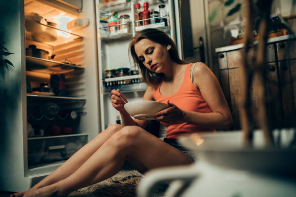 Woman in pajamas in front of the refrigerator late night Young woman wearing pajamas looking for a snack in the refrigerator late night eating disorder stock pictures, royalty-free photos & images