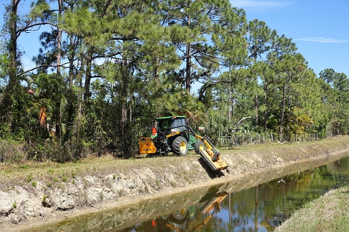A John Deere tractor with a large mowing attachment cuts grass and brush on a south Florida canal bank in Palm Beach county.