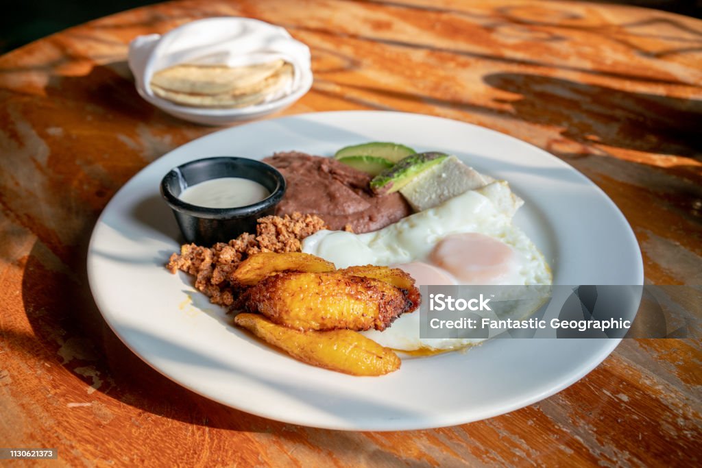 Traditional Honduran Breakfast with chorizo, fried eggs and plantains on a wooden table Honduras breakfast with fried plantains, eggs, beans, chorizo, crema and corn tortillas Honduras Stock Photo