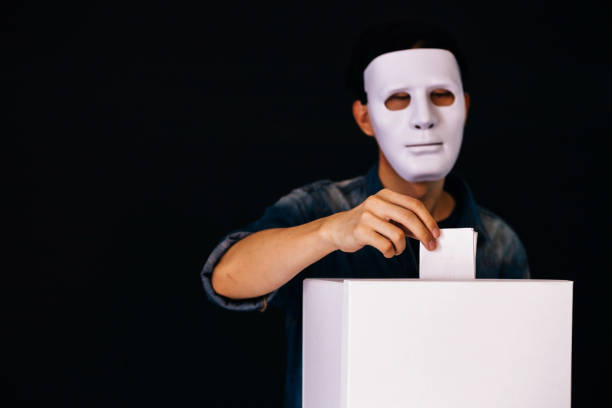 Masked criminal holding ballot paper casting fake vote at a polling station for election vote in black background. stock photo