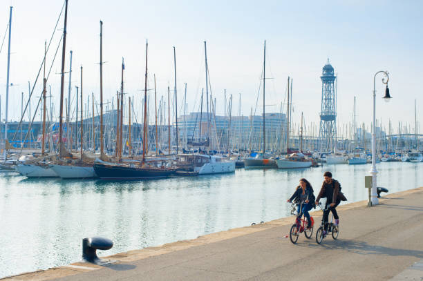 Couple riding bicycles by Barcelona marina with yachts, Spain