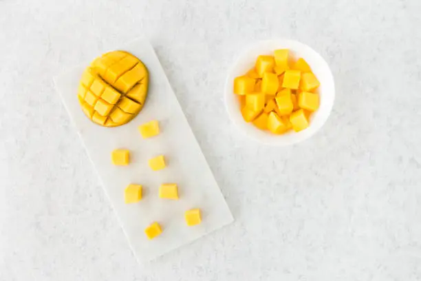 Top view of cut mango half and mango cubes on white marble cutting board and in white bowl.