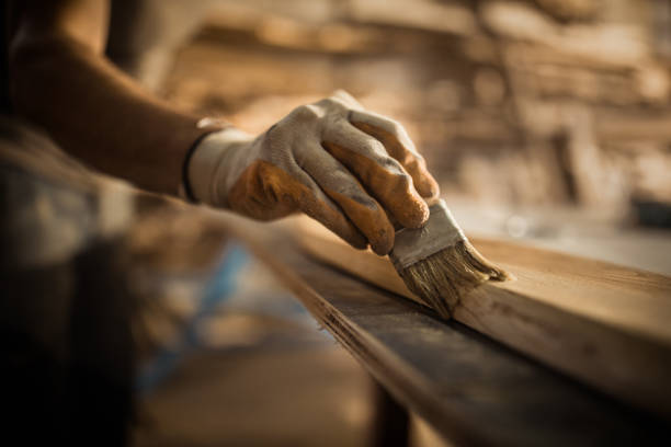 Close up of carpenter using brush while applying protective varnish to a piece of wood. Close up of carpenter applying varnish with a brush on a piece of wood in a workshop. wood stain stock pictures, royalty-free photos & images