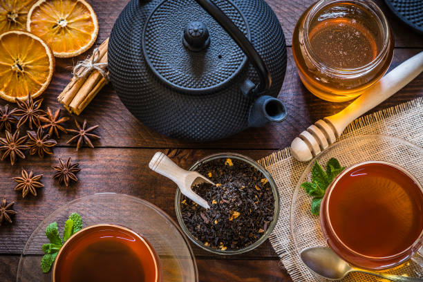 Tea time: cup of tea, cinnamon sticks, anise, dried orange on wooden table Top view of a rustic wooden table with two tea cups, a teapot and a crystal bowl filled with dried black tea, a honey jar with honey dipper and some ingredients like cinnamon sticks, anise and dried orange for preparing tasty tea. Low key DSLR photo taken with Canon EOS 6D Mark II and Canon EF 24-105 mm f/4L black tea stock pictures, royalty-free photos & images