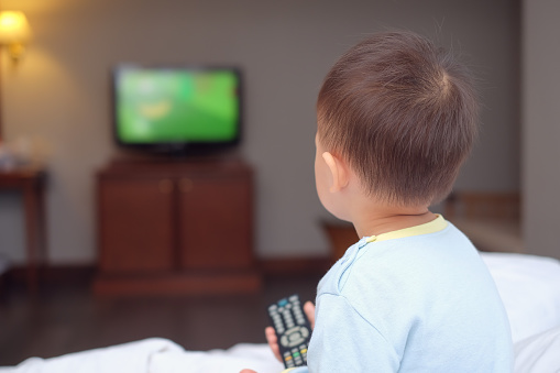 Lindo poco asiático 2-3 años de edad bebé niño niño sentado en la cama sosteniendo el control remoto de televisión y viendo televisión photo