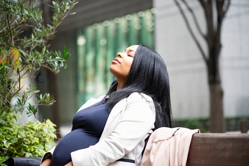 A mid adult pregnant woman sitting on a bench, enjoying the sunlight.