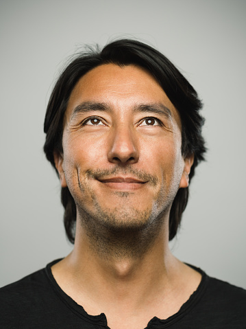 Close up portrait of adult hispanic man with happy expression looking up against white gray background. Vertical shot of colombian real people smiling in studio with black hair and dark eyes. Photography from a DSLR camera. Sharp focus on eyes.