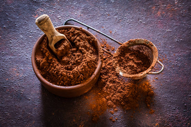 Top view of a brown bowl filled with cocoa powder shot on rustic brown table. Beside the bowl is a sieve also with cocoa powder. Predominant color is brown. Low key DSRL studio photo taken with Canon EOS 5D Mk II and Canon EF 100mm f/2.8L Macro IS USM.