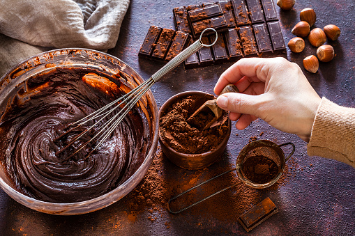 High angle view of a mixing bowl filled with melting chocolate dough shot on dark brown rustic kitchen table. A wire whisk is inside the mixing bowl. A female hand holding a wooden serving scoop is picking a cocoa powder from a small brown bowl. Some chocolate bar pieces are at the top right of an horizontal frame as well as some hazelnuts. A brown bowl with cocoa powder is beside the mixing bowl. Predominant color is brown. Low key DSRL studio photo taken with Canon EOS 5D Mk II and Canon EF 100mm f/2.8L Macro IS USM.