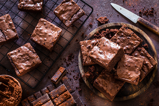 Top view of homemade chocolate brownies shot on dark table. A wooden plate filled with chocolate brownies is at the right and a cooling rack with brownies is at the left. Some chocolate bars are at the bottom. A brown bowl with cocoa powder is at the bottom left corner of an horizontal frame. Predominant color is brown. Low key DSRL studio photo taken with Canon EOS 5D Mk II and Canon EF 100mm f/2.8L Macro IS USM.