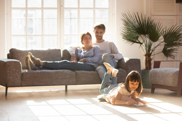 Happy parents relaxing on couch while kid drawing on floor Happy parents relaxing on couch in comfort light living room while little kid child daughter playing on warm floor drawing with colored pencils, family having fun together, underfloor heating concept family home stock pictures, royalty-free photos & images