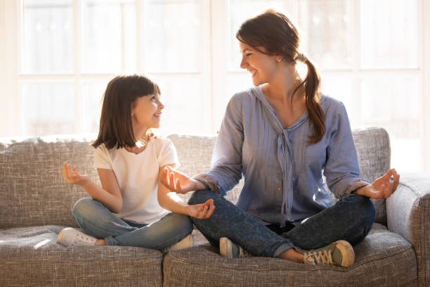 Mindful mom teaching yoga happy child daughter sitting on couch Mindful young mom teaching yoga happy cute funny child daughter sitting in lotus pose on couch together, smiling mother and little kid girl meditating relaxing at home, healthy family exercises mindfulness kids stock pictures, royalty-free photos & images