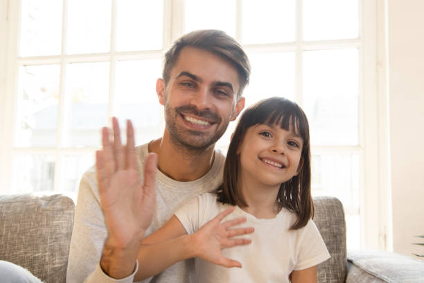 Happy cute little child girl and father youtubers vloggers wave hands say hello record vlog, smiling dad with kid daughter looking at camera webcam make online video call together at home, portrait