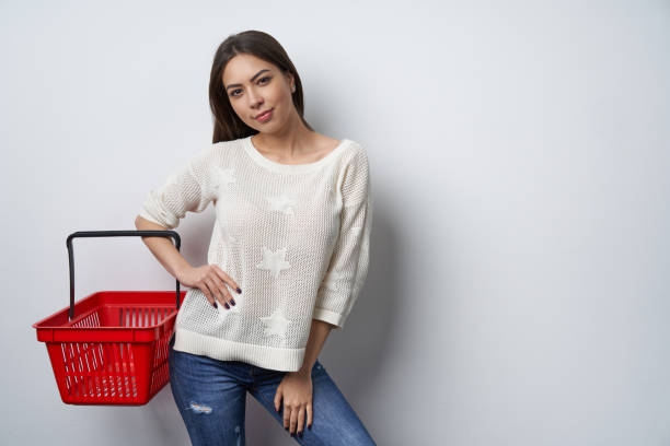 istock Smiling brunette woman standing by white wall holding empty shopping basket 1130701018