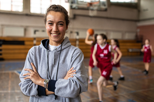 Young beautiful female basketball coach standing in school gym with crossed arms , looking at camera and smiling